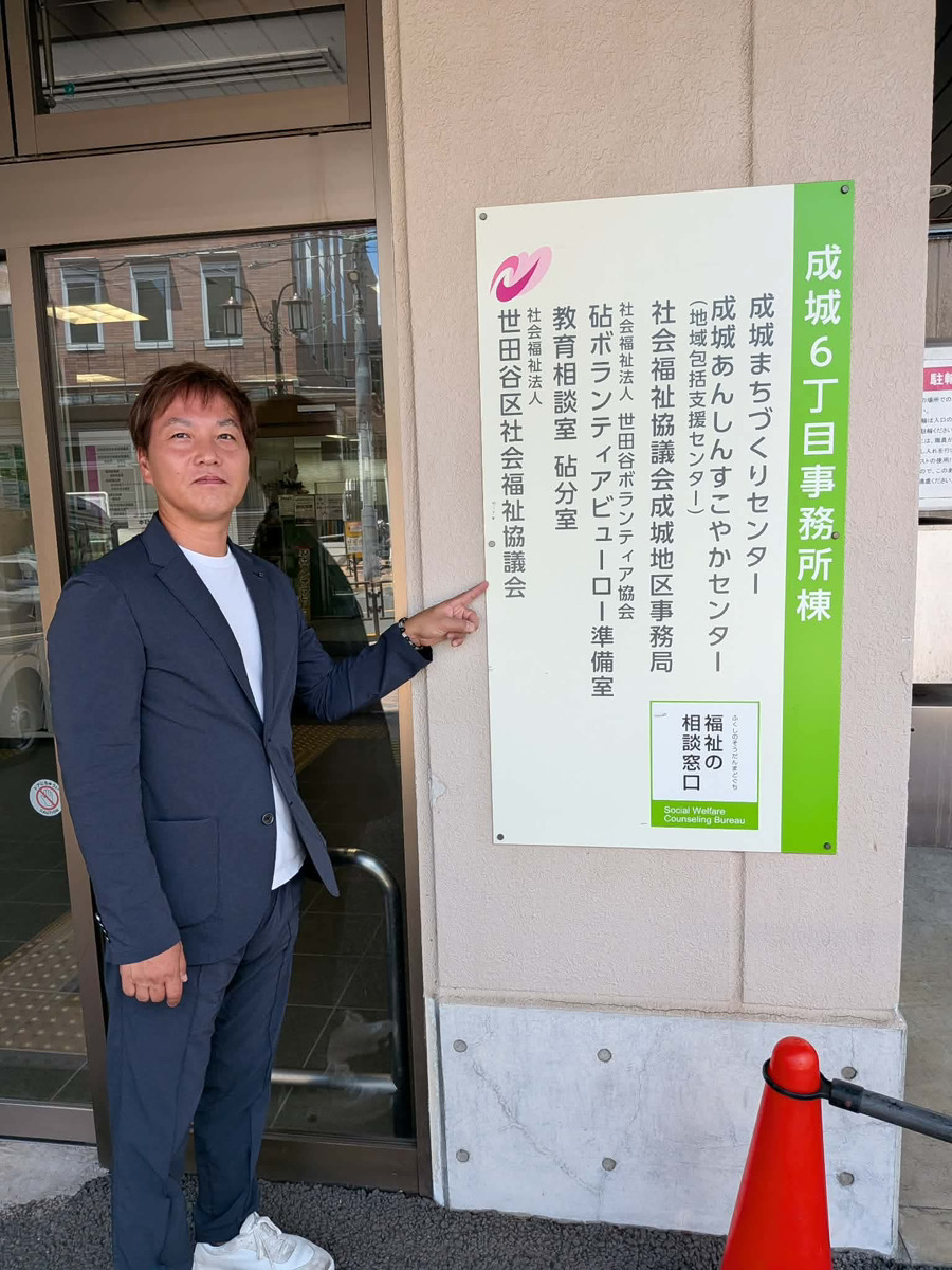 成城6丁目事務所棟の建物外観と入口看板を指差すガヤ経理事メンバー